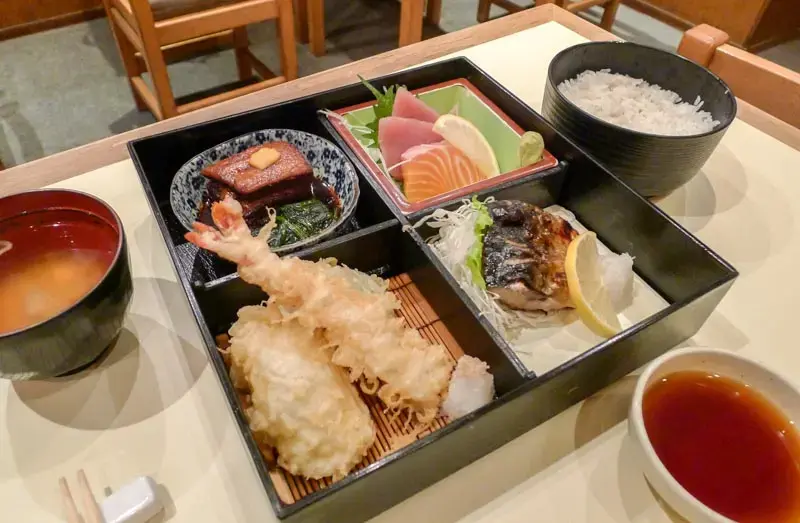 This image features a traditional Japanese bento box set served at a restaurant, containing assorted components like sashimi, tempura, grilled fish, and braised pork. The meal is accompanied by a bowl of white rice, a side of miso soup, and a small dish of dipping sauce on a light-colored table.