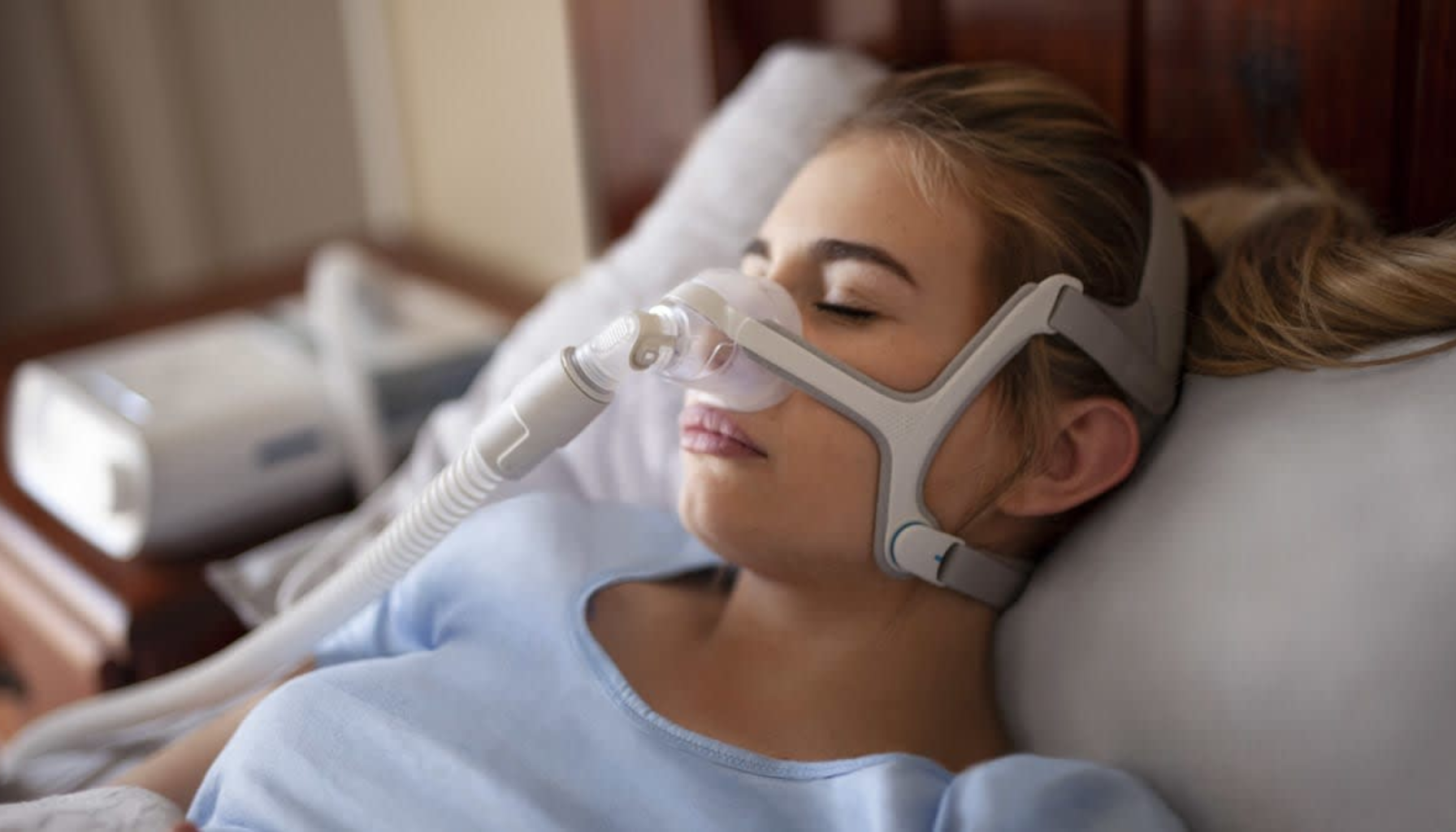 Ung kvinne sover med CPAP-maske og pusteslange festet, liggende i seng i rolige omgivelser.