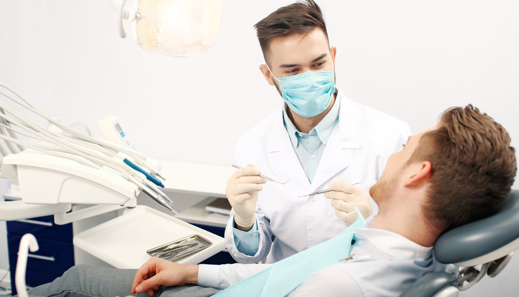 Tannlege med munnbind snakker med mannlig pasient i tannlegestol før undersøkelse i en moderne klinikk.