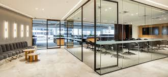 A sleek and modern office space featuring a glass-walled conference room. The conference room has a long table surrounded by black ergonomic chairs and is equipped with modern amenities. Outside the conference room, there is a spacious waiting area with a row of grey cushioned chairs and small wooden side tables. The office is well-lit with recessed lighting and has a neutral color scheme with light beige floors and walls. Large windows in the background allow natural light to illuminate the space, creating a bright and professional atmosphere. The design emphasizes transparency, openness, and functionality.