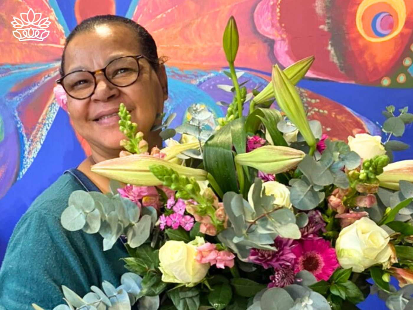 A delighted florist holding a pastel flower bouquet for secretaries delivery featuring white roses, pink snapdragons, and green eucalyptus leaves, complemented by vibrant artistic mural backdrop, from the Flowers By Occasion Collection at Fabulous Flowers and Gifts.