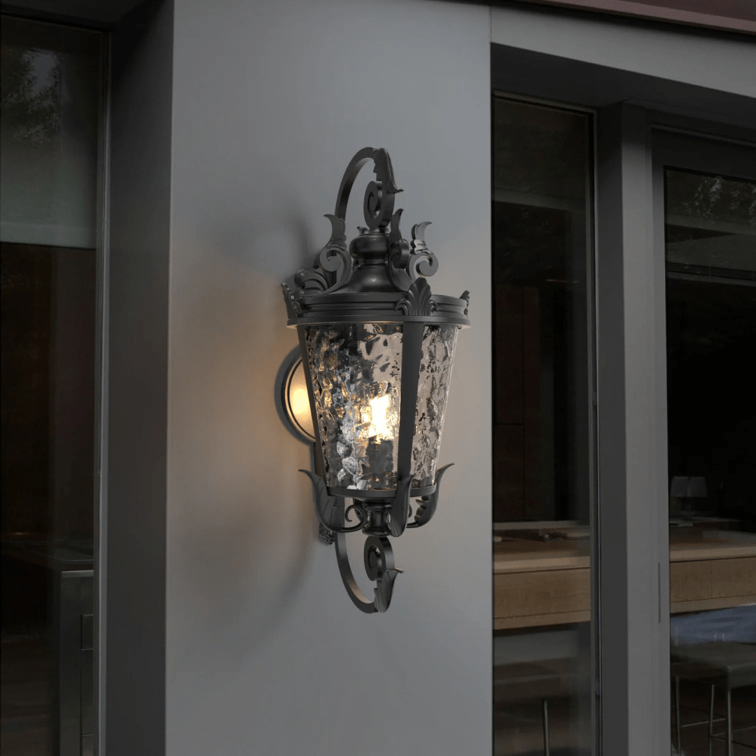 Ornate black outdoor wall lantern with decorative scrollwork and textured glass panels, mounted on a modern gray wall.
