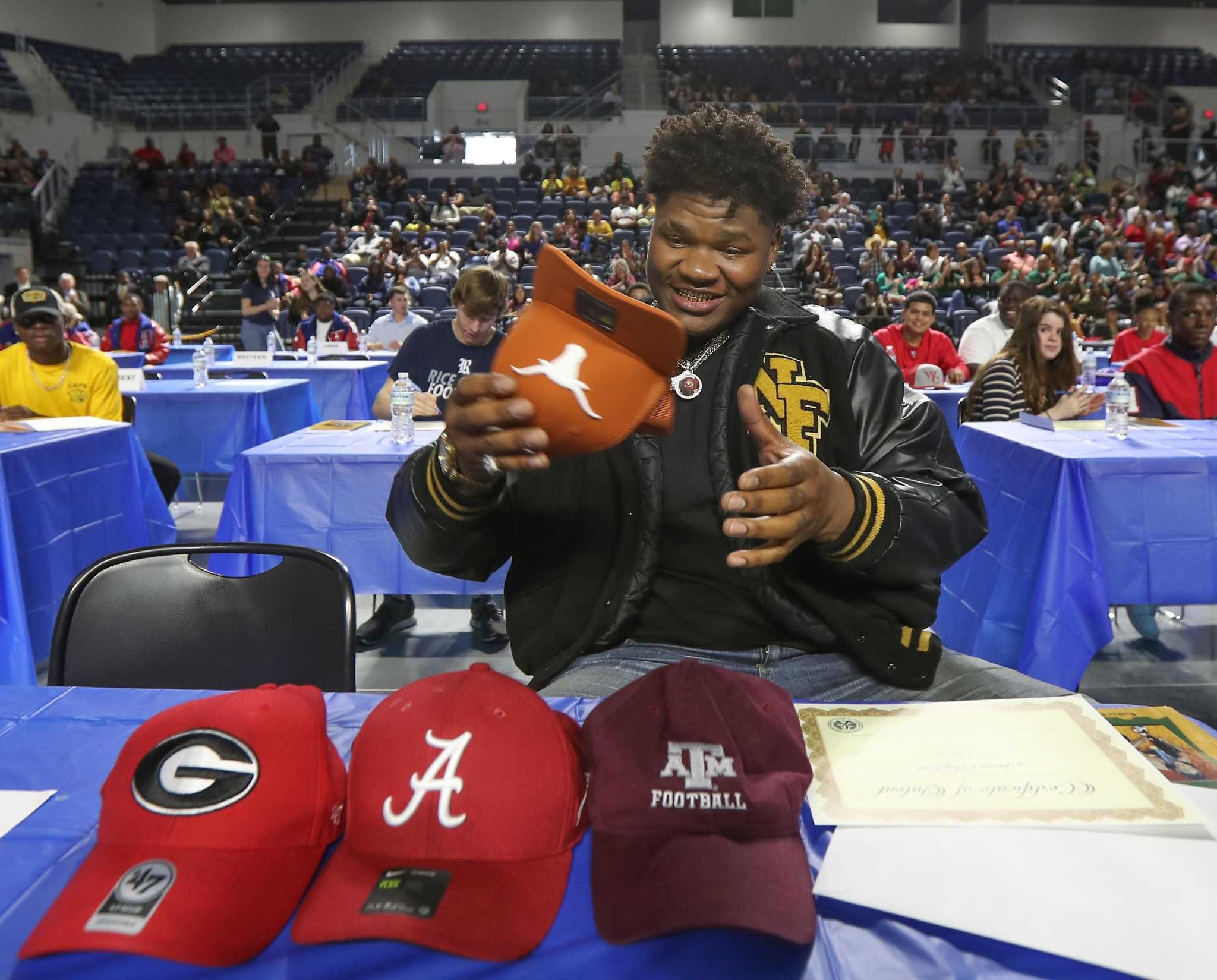 Student-athlete at a signing ceremony grins while picking up a burnt orange Texas Longhorns hat from a table lined with other college hats, including Georgia, Alabama, and Texas A&M. The event is held in a large gym with other students and families in the background.