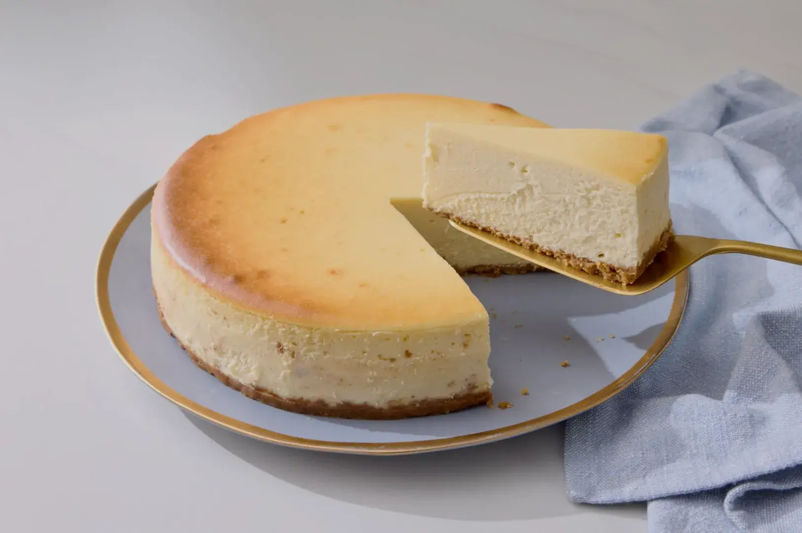 A round cheesecake on a plate, with a slice being lifted out by a serving utensil. A light blue cloth is partially visible next to the plate.
