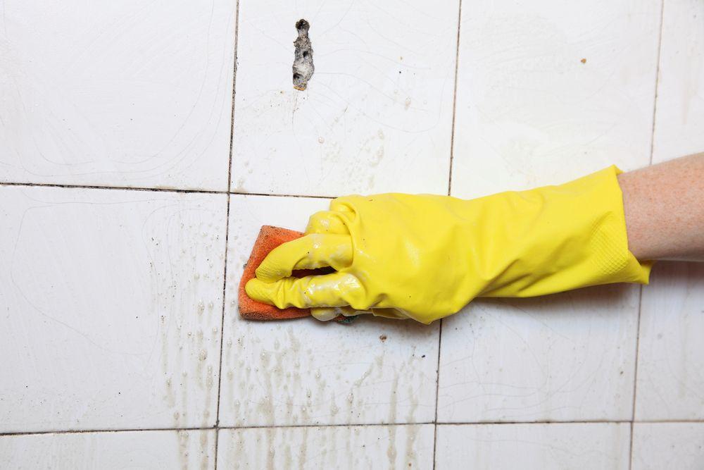 Person wearing yellow gloves scrubbing dirty tile and grout with a cleaning sponge.