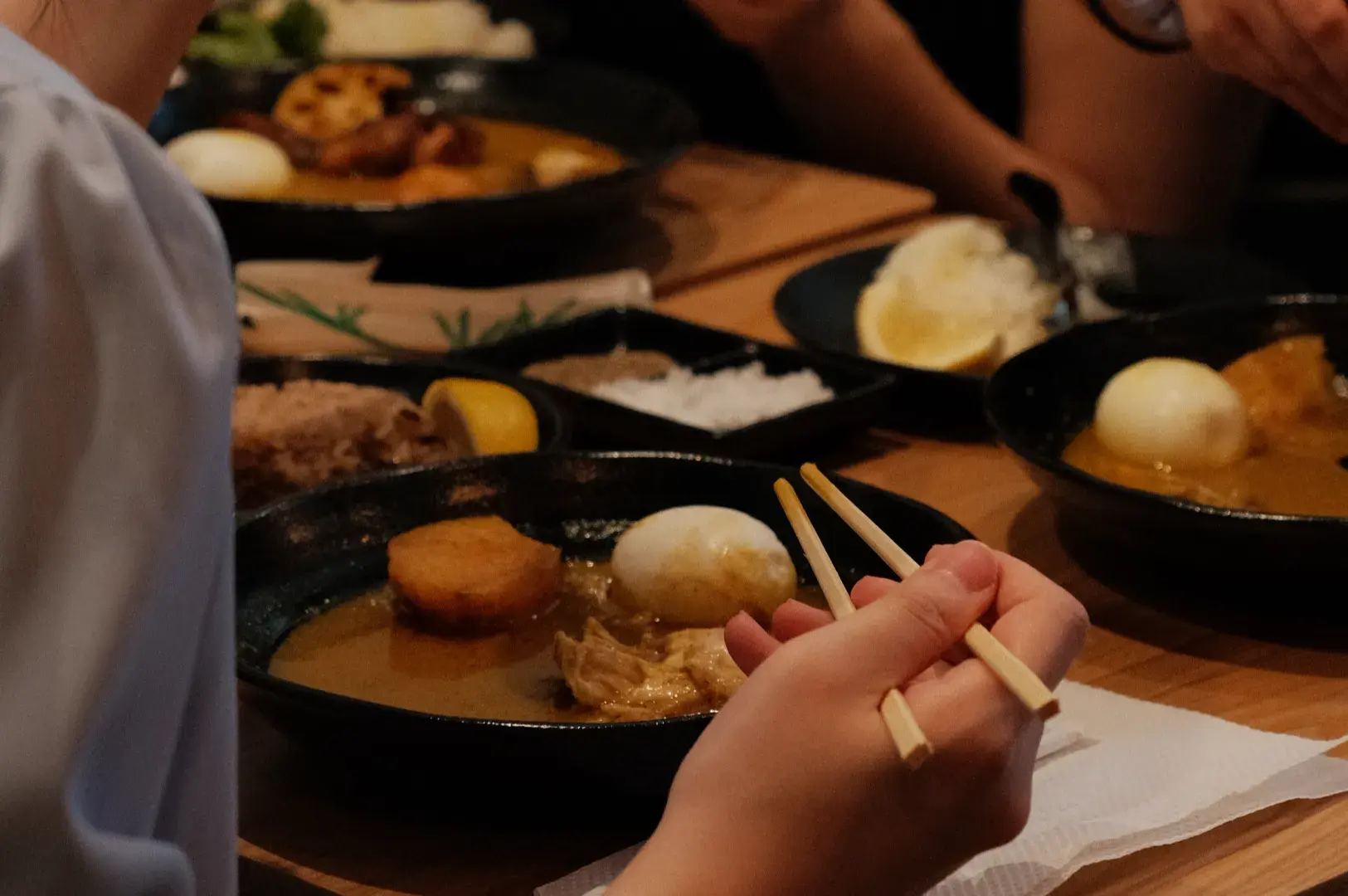 A close-up of hands using chopsticks to eat a bowl of curry with an egg and meat, with plates of food and other diners in the background.