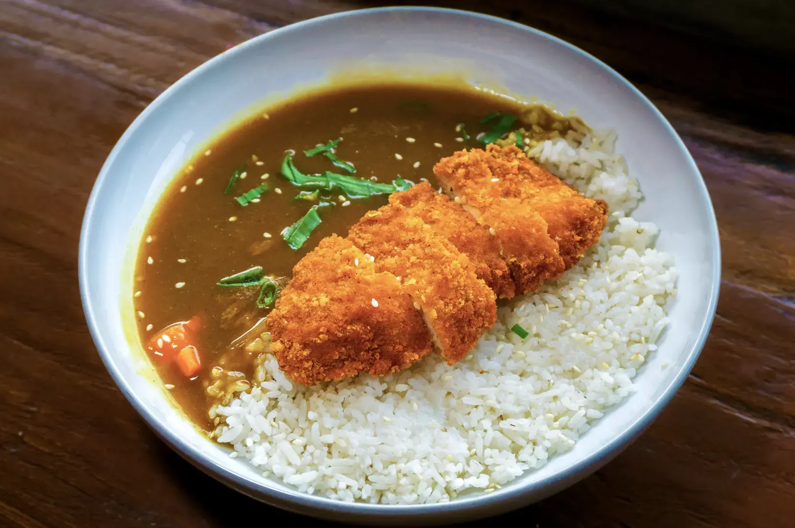 A white bowl of Japanese curry with rice topped with sliced katsu chicken, garnished with sesame seeds and green onions.