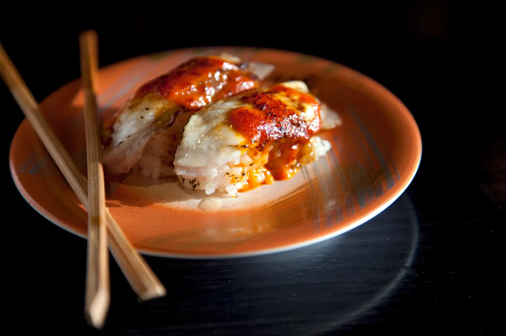 Two pieces of torched, glazed Aburi nigiri sushi served on an orange plate with chopsticks.