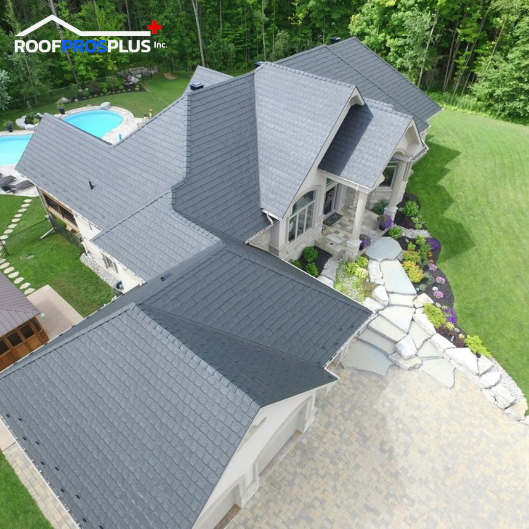 An aerial view of a large house with a dark, textured metal roof surrounded by lush green lawn and forest. A stone path leads to a pool area.