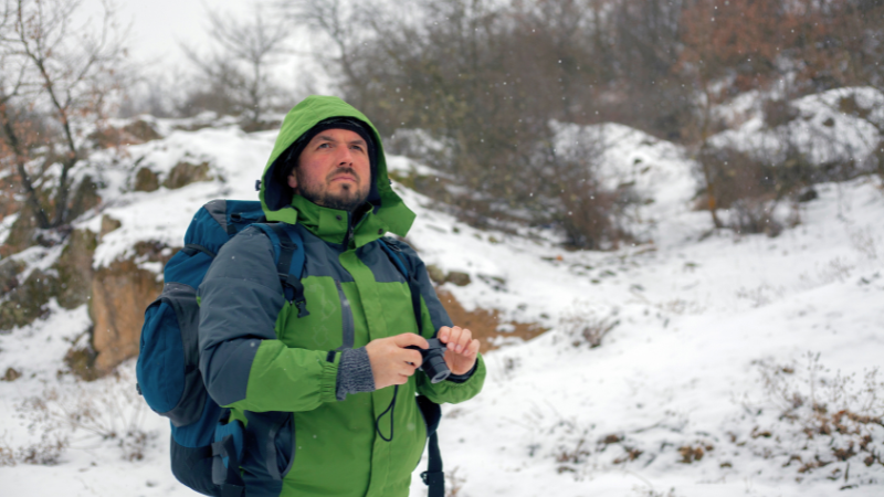 A person in a green jacket and backpack uses binoculars in a snowy landscape.