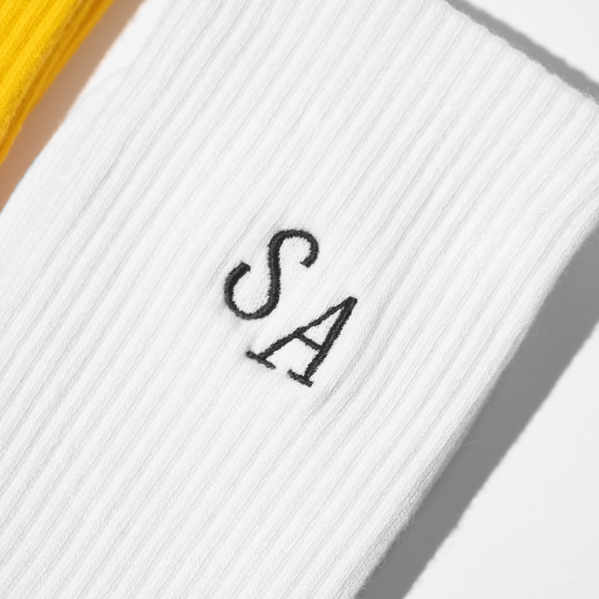A flat-lay image of custom mens white crew socks with the initials 'SA' embroidered in black thread.