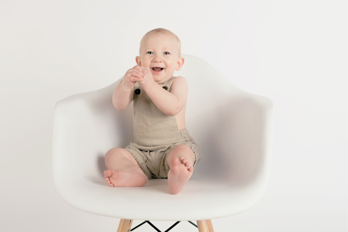 Baby boy sitting up on white chair. Poppyseed Play