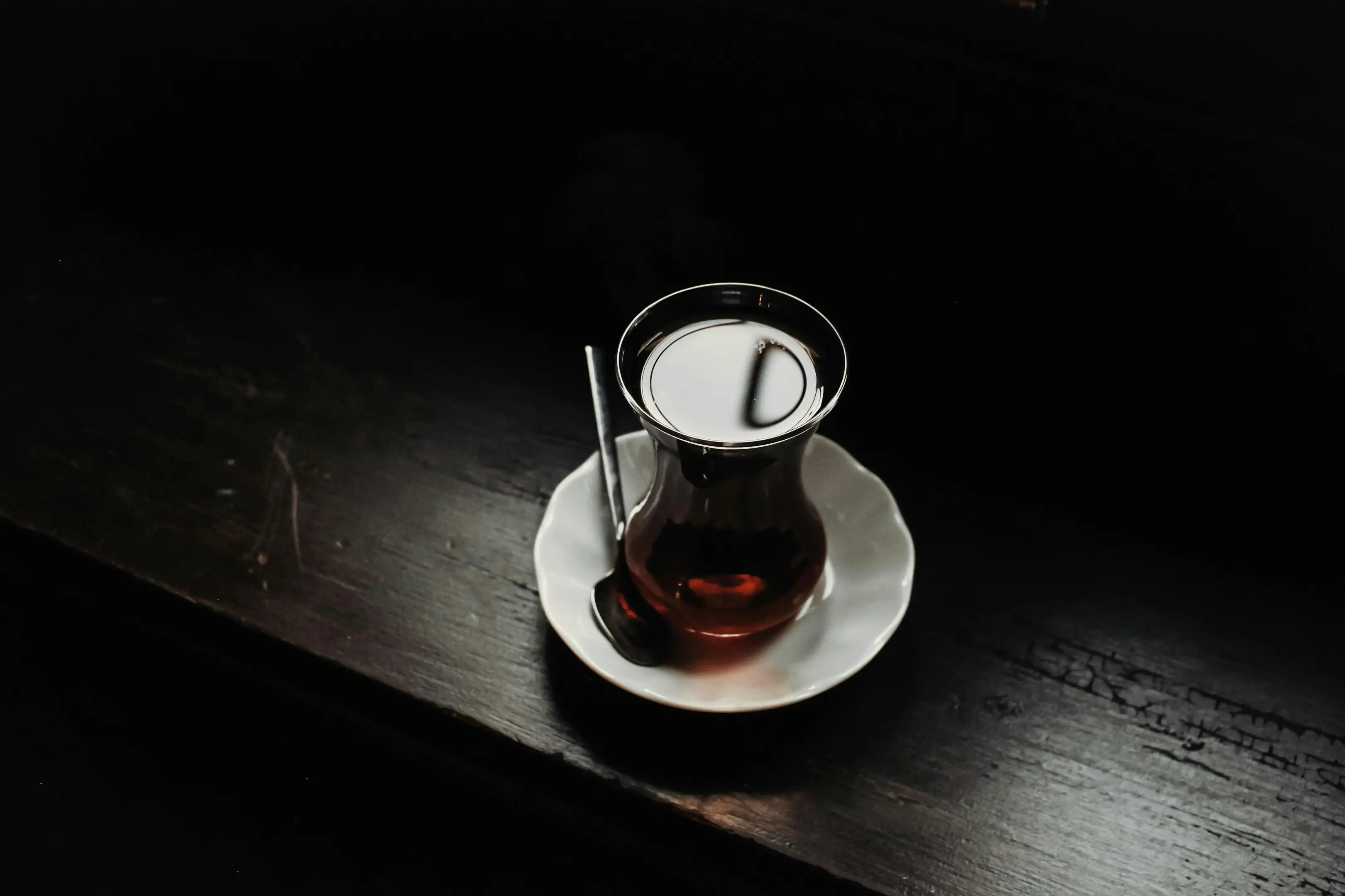 A small, tulip-shaped glass filled with dark tea rests on a white saucer with a small spoon, positioned on a dark wooden surface. The moody, low-light setting creates a dramatic contrast, highlighting the liquid's surface reflection and warm amber hue.