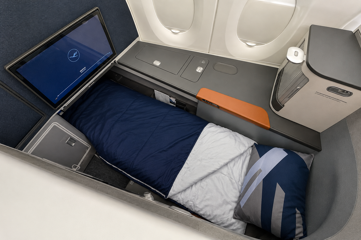 Business class suite with a fully flat bed, navy duvet, white bedding, geometric pillow, entertainment screen and storage beside aircraft windows.