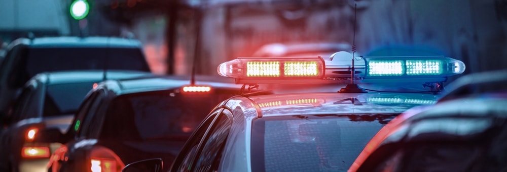 A police officer standing next to a stopped vehicle with flashing lights visible in the background.