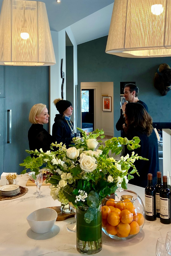 Small group of adults conversing in a modern kitchen with pendant lighting, floral centerpiece, wine bottles, and bowls of citrus on a marble island.