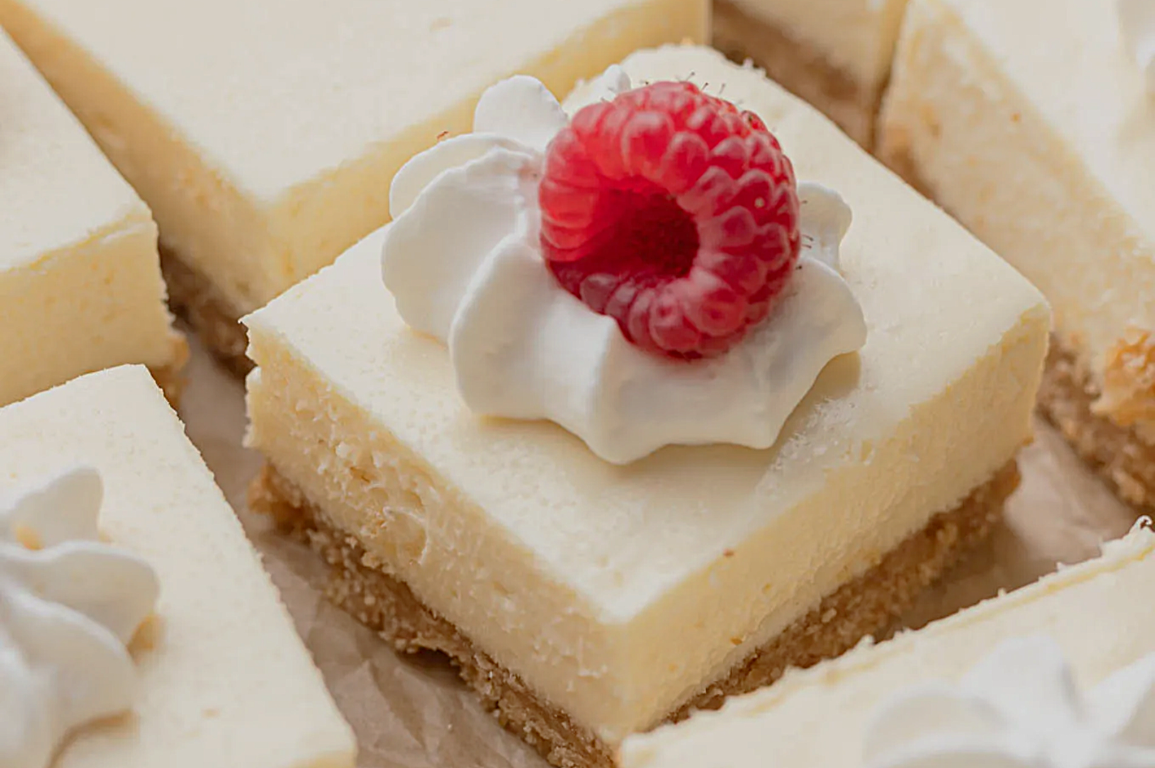A creamy cheesecake square topped with whipped cream and a fresh raspberry, resting on a crumbly crust, conveys a sense of elegance and indulgence.