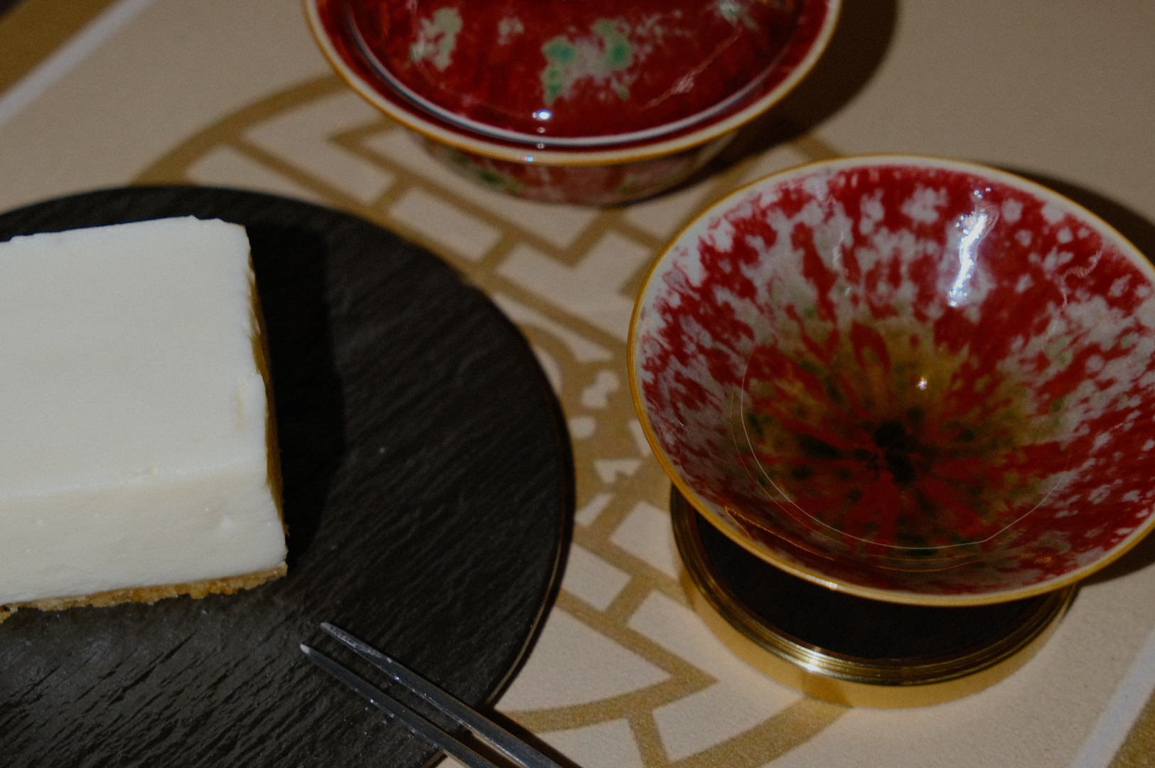 A slice of cheesecake sits on a black plate beside two red and white ceramic bowls with intricate patterns, on a patterned beige surface.