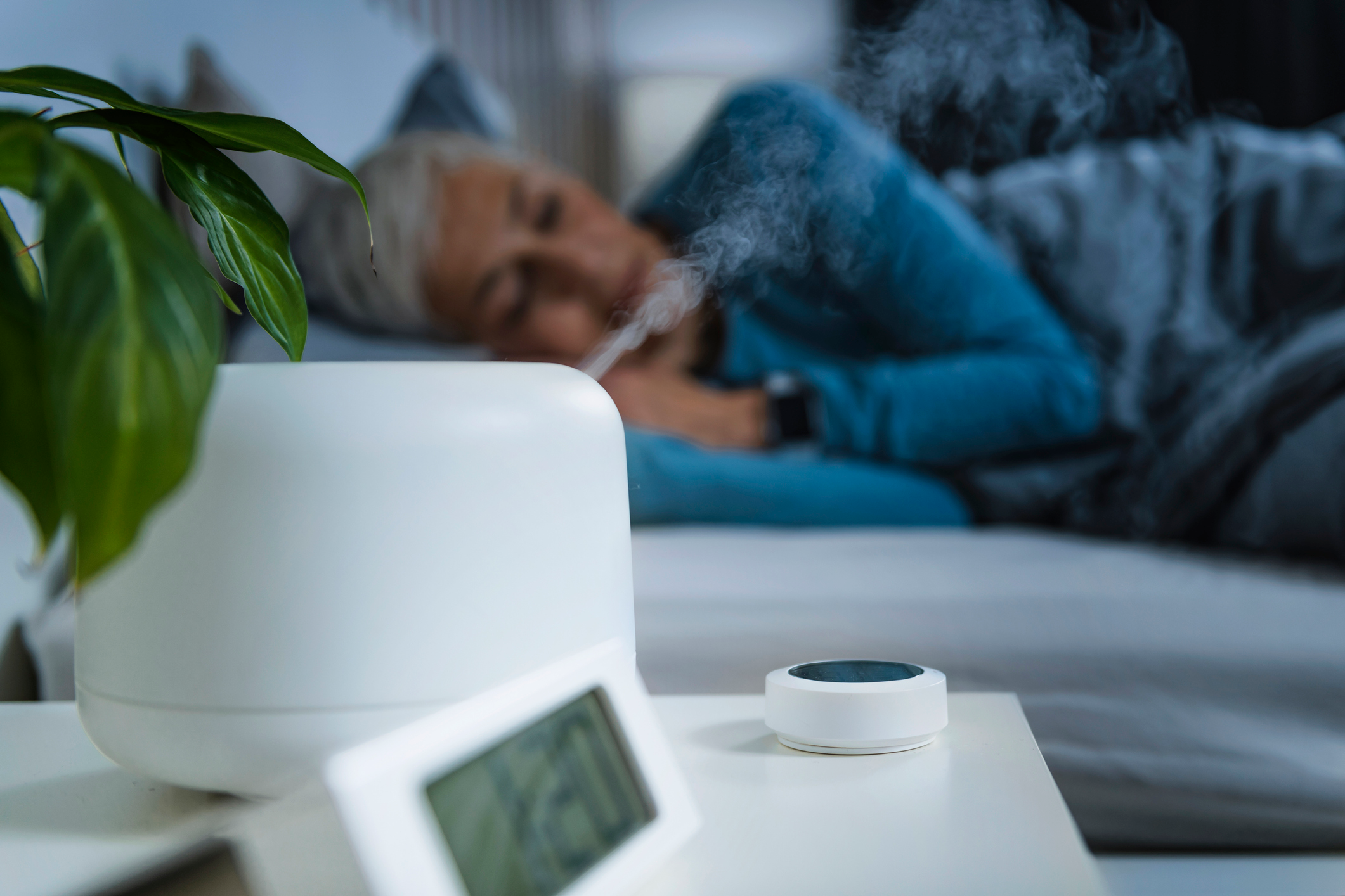 An image of a mature woman sleeping in bed breathing steam from the humidifier on the night table. 
