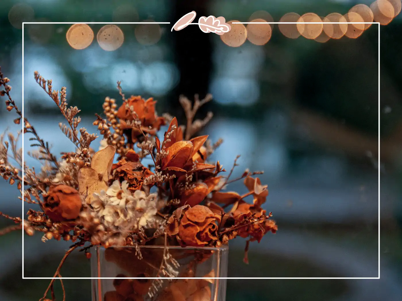 A charming arrangement of dried flowers in a glass vase, including orange and white blooms, set against a backdrop of twinkling bokeh lights, creating a cozy and enchanting ambiance. Fabulous Flowers and Gifts.