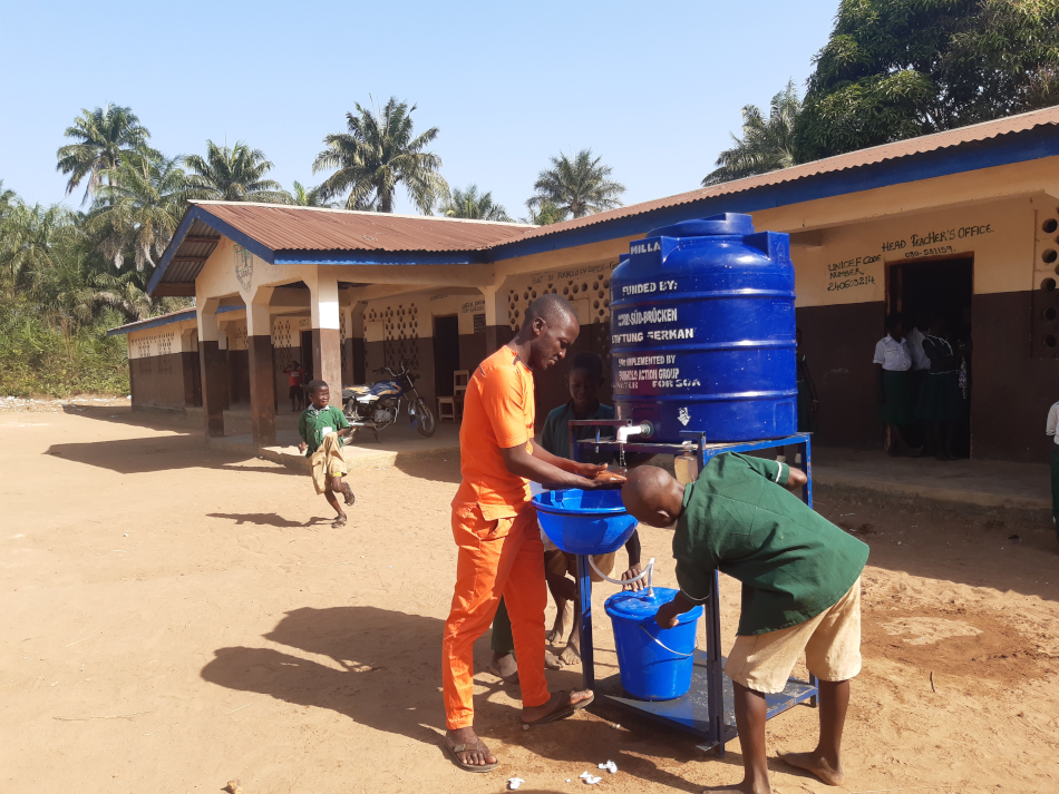 Aufnahme einer Handwaschstation, die von einem Lehrer und zwei Schülern genutzt wird, vor einer Schule in Sierra Leone. Erfahre alles zum Gesundheitssystem in Sierra Leone!