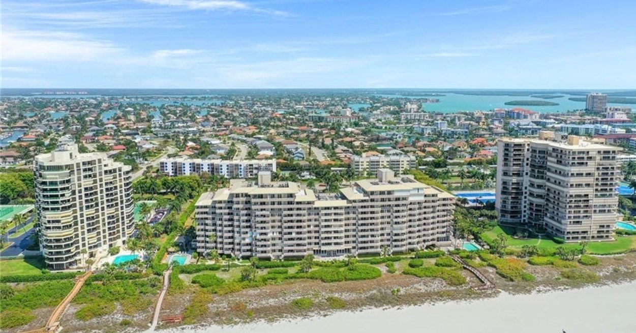 The image depicts a luxurious high-rise condominium building overlooking a marina filled with boats, embodying the essence of island living in Marco Island, Florida. This stunning property offers breathtaking views and easy access to the beach, making it an ideal choice for those seeking luxury living on the Paradise Coast.