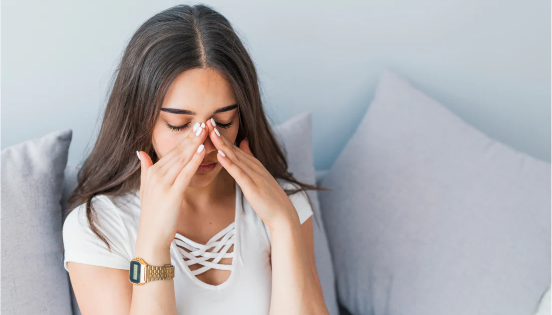 Kvinne som sitter på sofa og masserer neseroten med øynene lukket, sannsynligvis ved bihulebetennelse.
