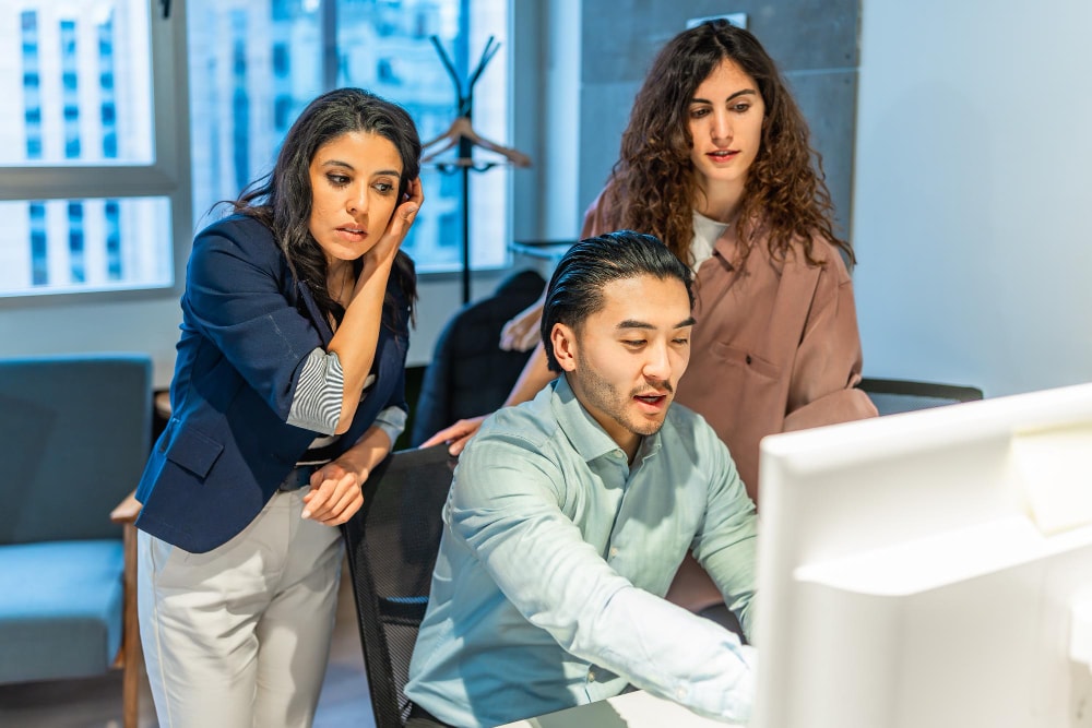 Three coworkers collaborating and discussing something on a desktop monitor in a corporate office, emphasizing teamwork and streamlined business communication through integrated phone system upgrades.