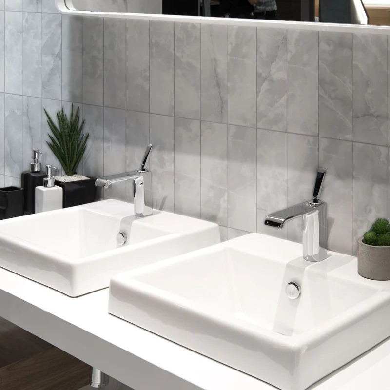 Vertically stacked glossy grey marble-effect wall tiles behind twin square white basins with chrome taps and minimalist decor.