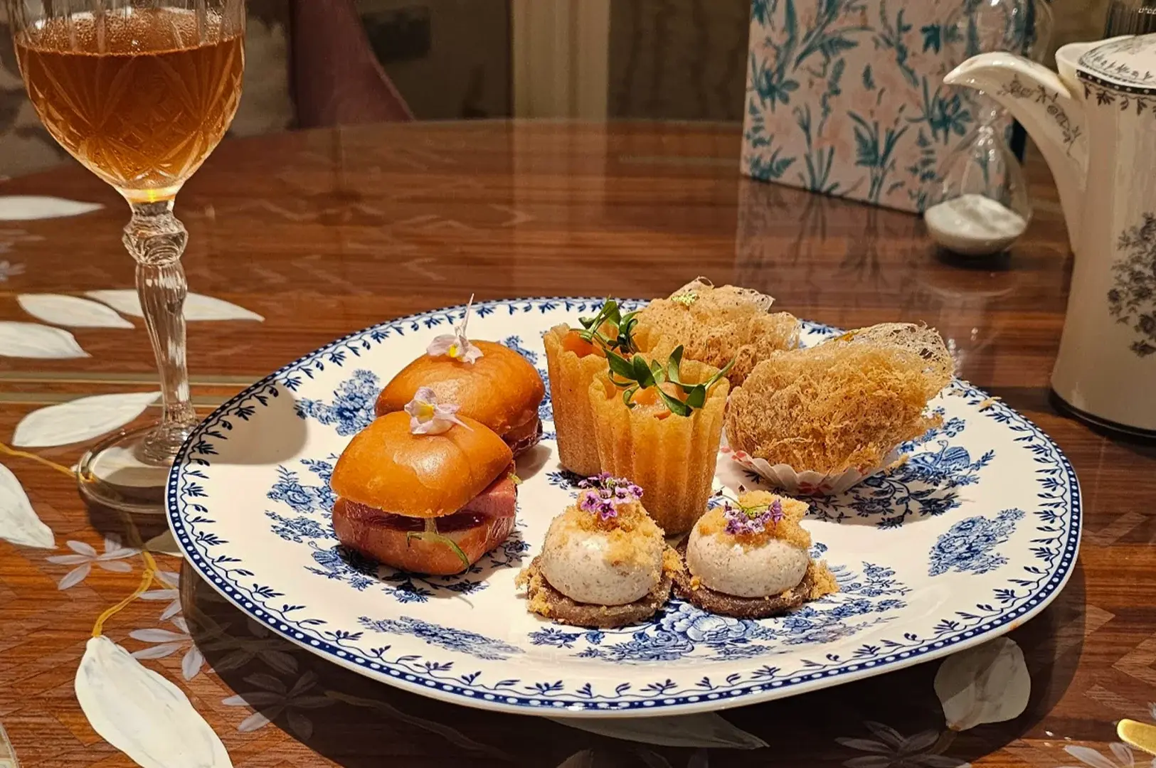 A close-up photograph showcases a selection of savory afternoon tea delicacies beautifully arranged on a white porcelain plate with an intricate blue floral border, sitting on a polished dark wood table. The plate features two glossy, golden-brown slider buns garnished with tiny purple orchids, two crispy cylindrical pastry cups filled with a salmon mixture and microgreens, two golden, shredded bird's nest-style fried pastries, and two small round crackers topped with a creamy mousse and micro-flowers. In the softly blurred background, a crystal glass filled with rose champagne stands beside a matching blue-and-white floral teapot and a white sand hourglass timer.