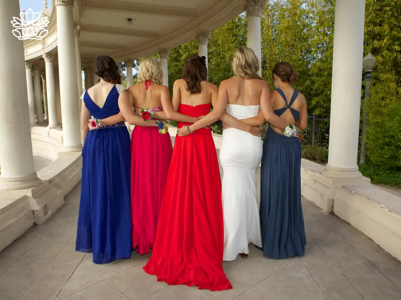 Rear view of five young women at a matric dance, arm in arm under a columned pavilion, each adorned in vibrant formal gowns of red, white, and blue and matching floral wrist corsages. Fabulous Flowers and Gifts - Matric Dance. Delivered with Heart.