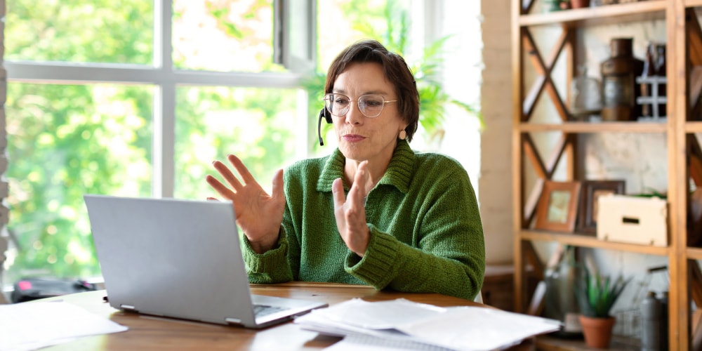 A woman works in front of a laptop monitor video conferencing and providing phone support.