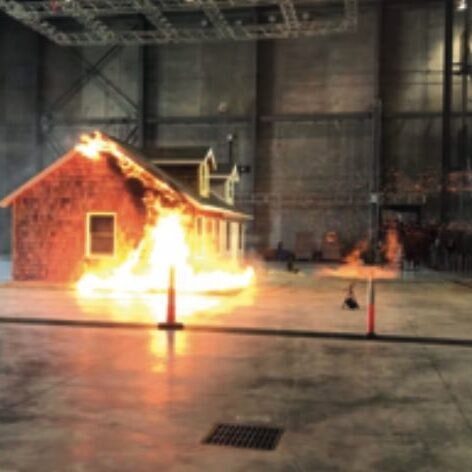 A picture of a metal roof being fire tested in a controlled environment. The demonstration is taking place in a large warehouse.