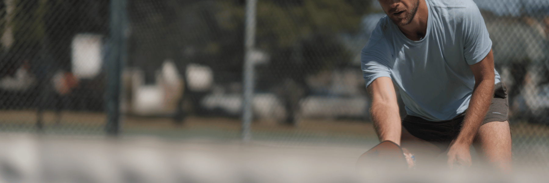 A man in an athletic stance leans forward on a pickleball court, seen from behind the net.
