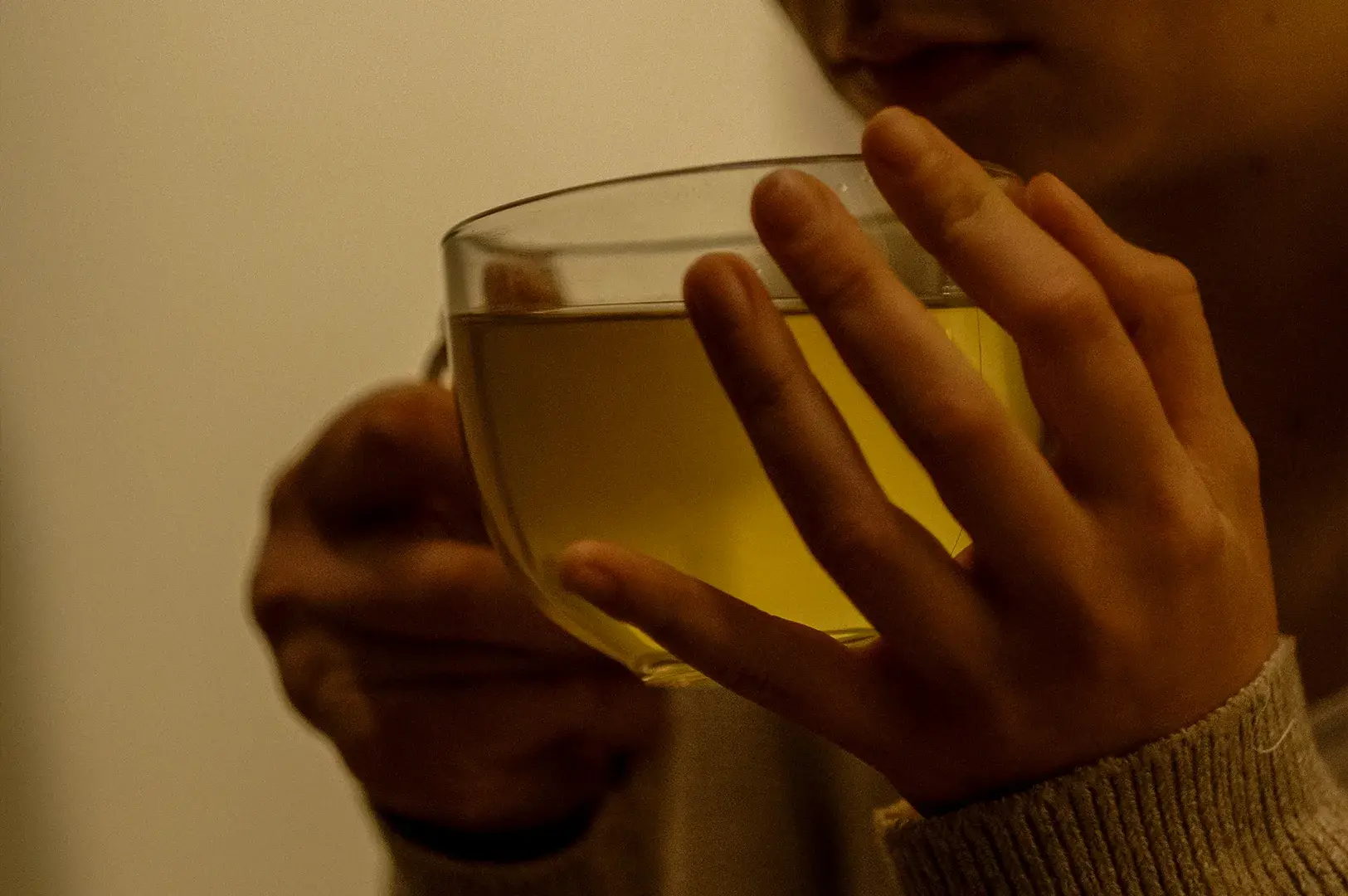 A warm, close-up shot of a person holding a large, clear glass mug filled with a pale golden-green tea up to their face, preparing to take a sip. The person's hands are wrapped around the body of the glass, showing the silhouette of their fingers against the warm translucent liquid, while they wear a cozy, ribbed knit sweater sleeve. The lighting is soft and dim, creating an intimate, comforting atmosphere focused on the simple act of enjoying a warm beverage.