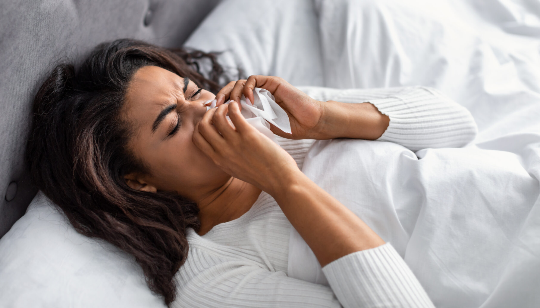 Kvinne ligger i seng og tørker nesen med papirserviett, tydelig plaget av forkjølelse eller allergi.