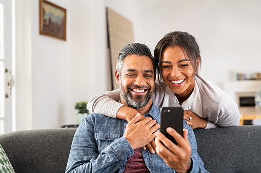 Pretty dark haired woman hugging a man as he sits on the sofa smiling at his cell phone. 