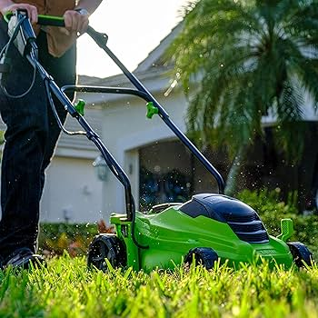 A person is using a green lawn mower to cut grass in a sunny, residential area with palm trees and a house in the background. The mower is close to the ground, capturing the moment of cutting grass blades.