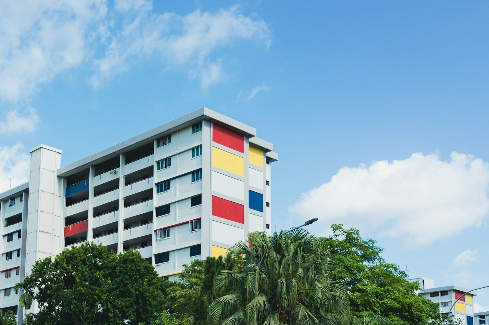 Colorful high-rise building with red, yellow, and blue panels, surrounded by lush green trees under a clear blue sky with clouds. Calm and vibrant urban scene.