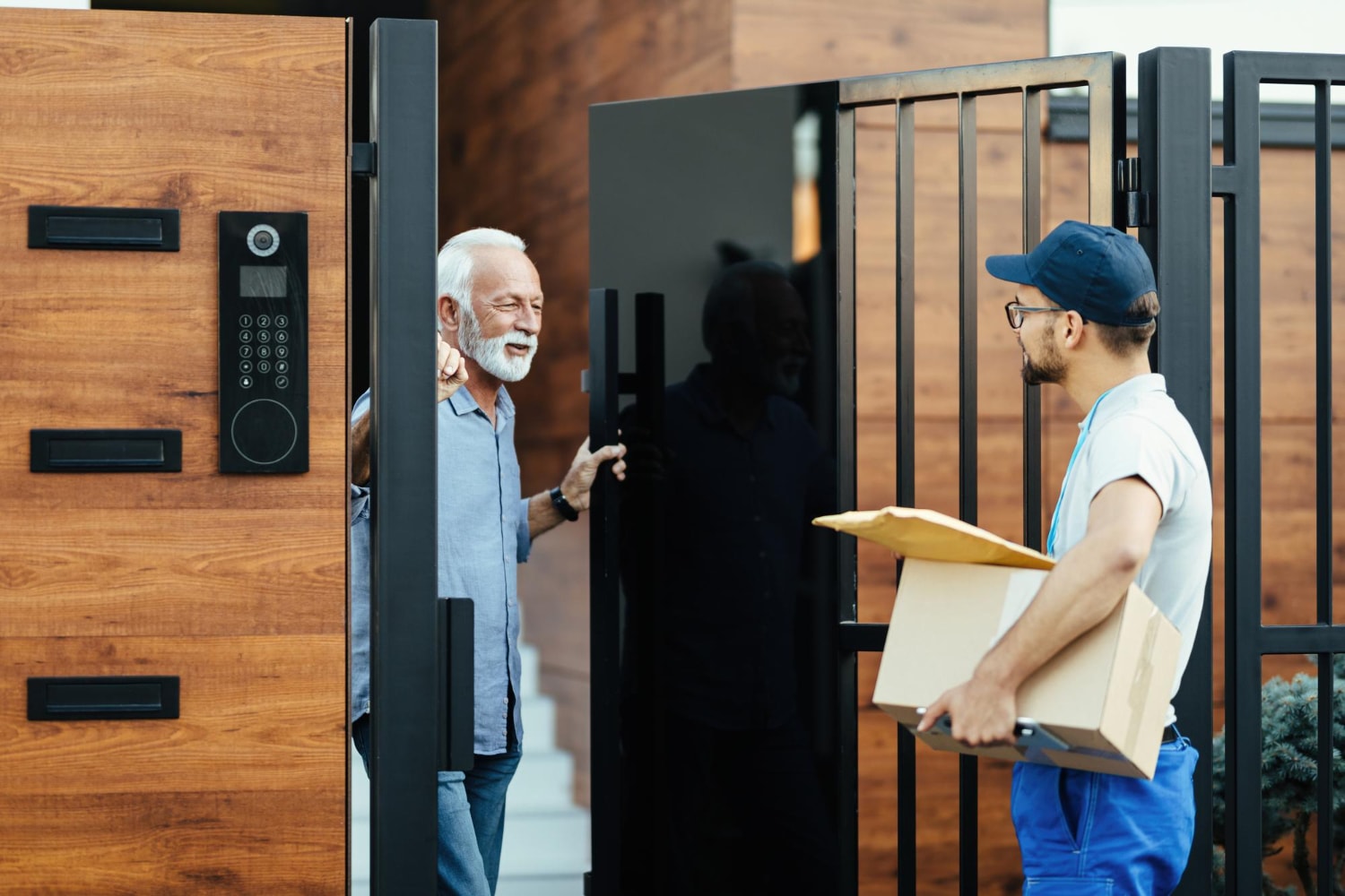An older man opens a gate with a built-in video intercom system to greet a delivery person holding packages. This residential access control scene highlights real-time visitor interaction via video intercom solutions, enhancing smart building security.