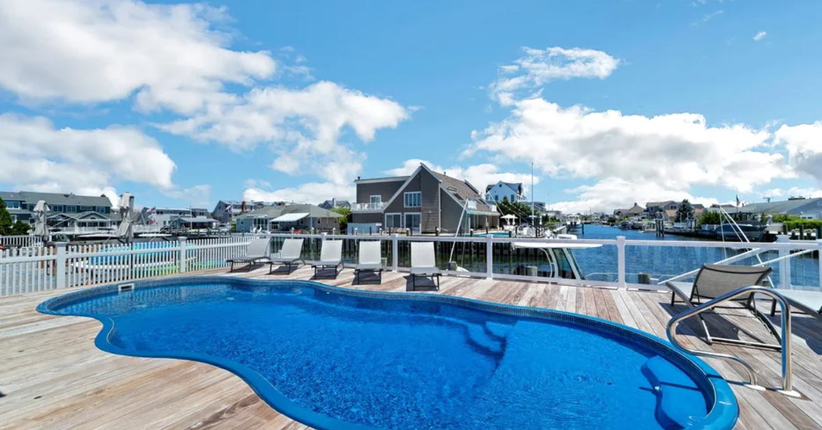 Backyard pool overlooking a peaceful Mantoloking waterfront with lounge chairs and coastal homes under a bright blue sky.