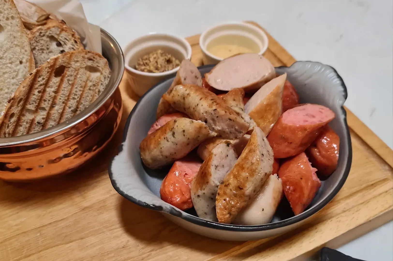 A rustic serving board holds a savory selection for the high tea, featuring a copper-toned pot filled with slices of toasted artisan bread and a small grey ceramic bowl overflowing with a variety of sliced, pan-seared sausages. Two small white ramekins containing dipping sauces—one yellow and one textured brown—are placed alongside the sausages, offering an appetizing and savory contrast to the traditional sweet offerings.