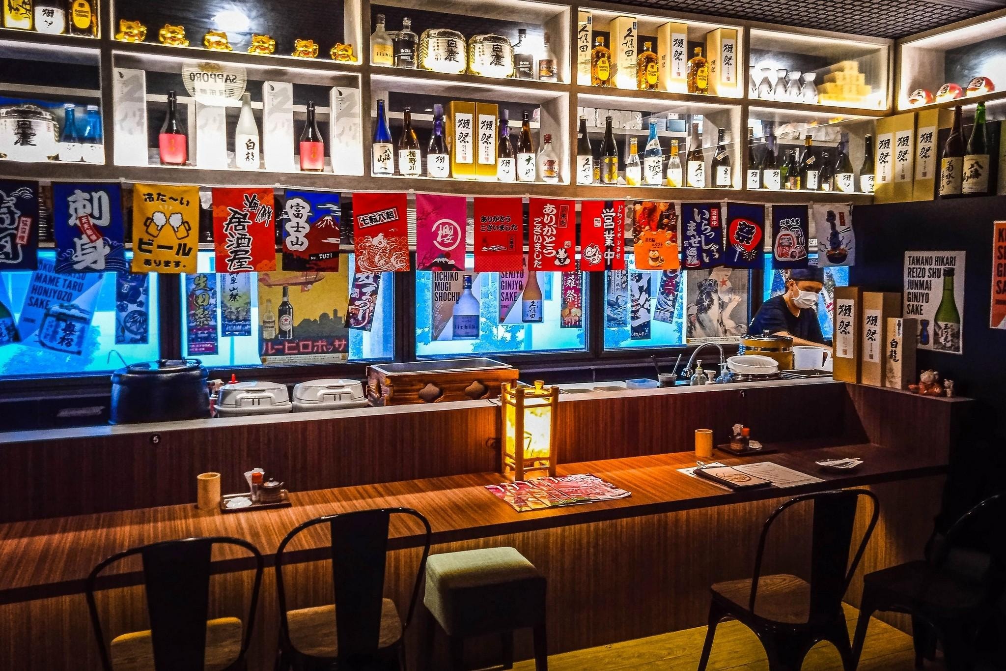This image showcases the warm, cluttered interior of Izakaya Hikari, a Japanese bar in Singapore, featuring a long wooden counter lined with dark metal chairs. Above the bar, shelves are packed with an extensive collection of sake bottles and traditional decor, while colorful Japanese banners hang across the service window.