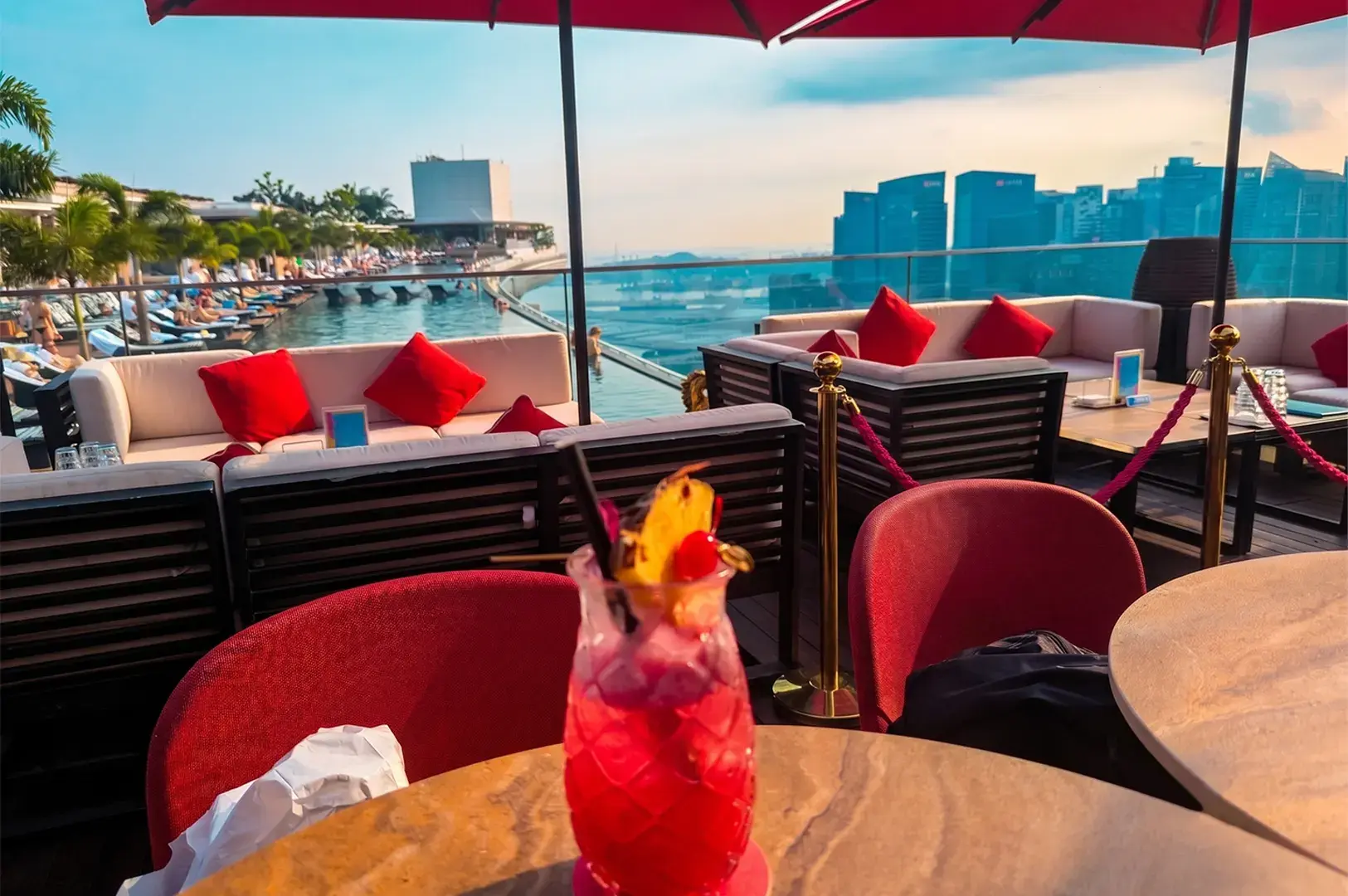 A refreshing red tropical cocktail served in a pineapple-shaped glass on a rooftop bar overlooking the Singapore skyline and infinity pool.