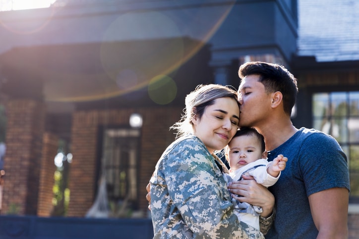 Husband kissing his soldier wife as she holds their baby. 