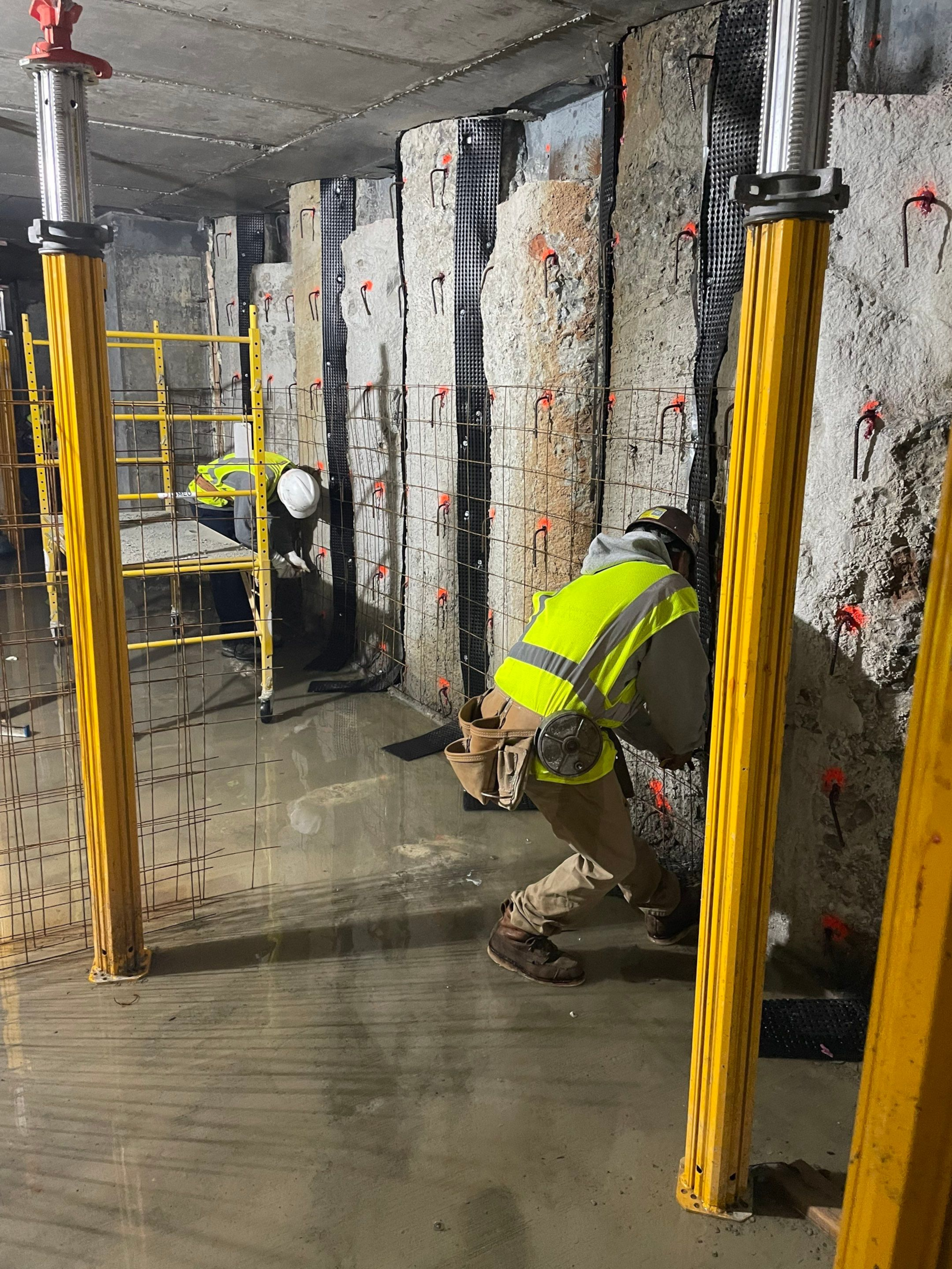 A contractor and his team applying shotcrete to a retaining wall at a construction site, showcasing the spraying process and steel reinforcement.