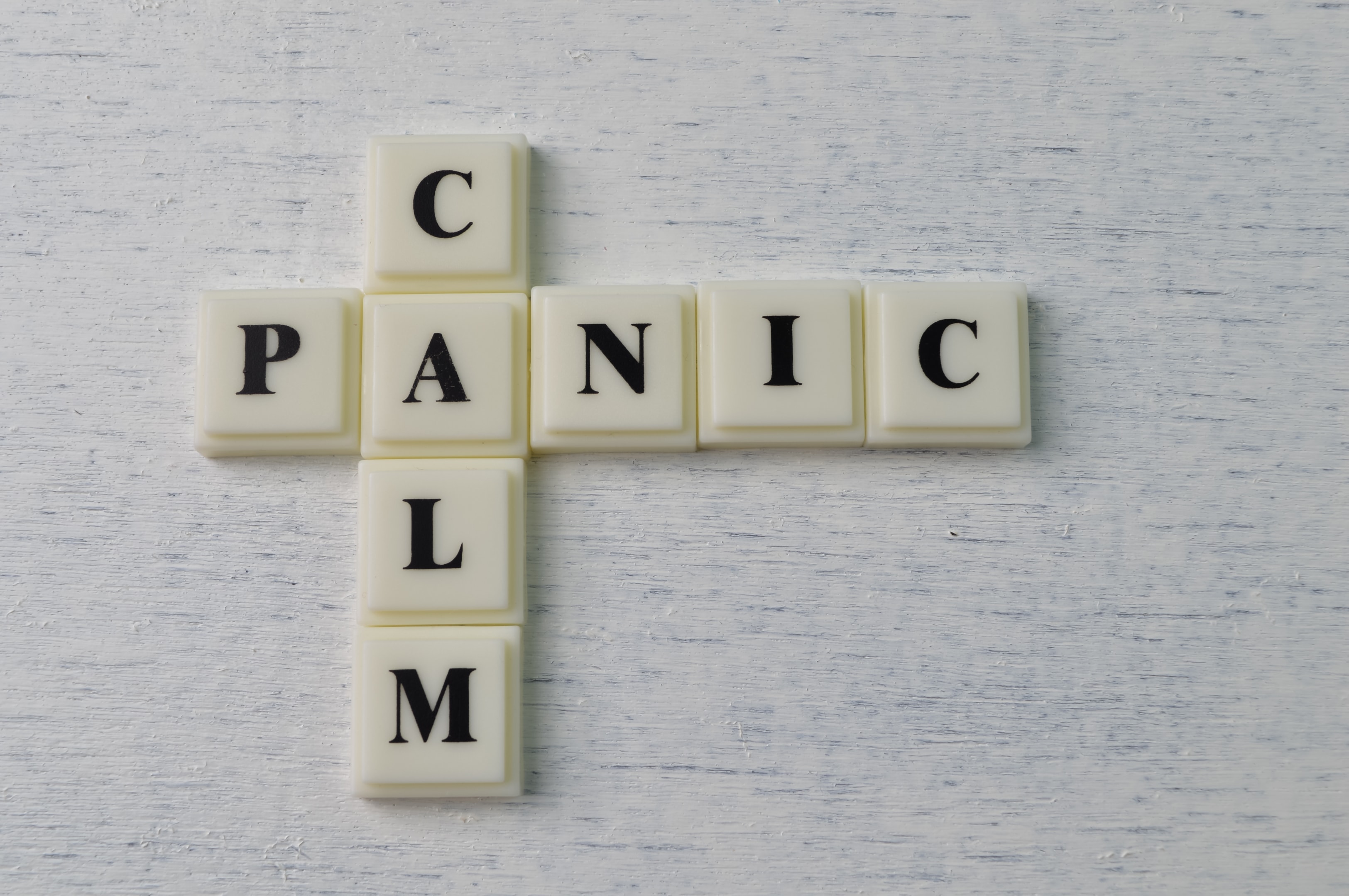 White game tiles arranged to form the word "PANIC" horizontally and "CALM" vertically on a light-colored wooden surface, representing the transition that Managing Panic Attacks in The Woodlands can facilitate.