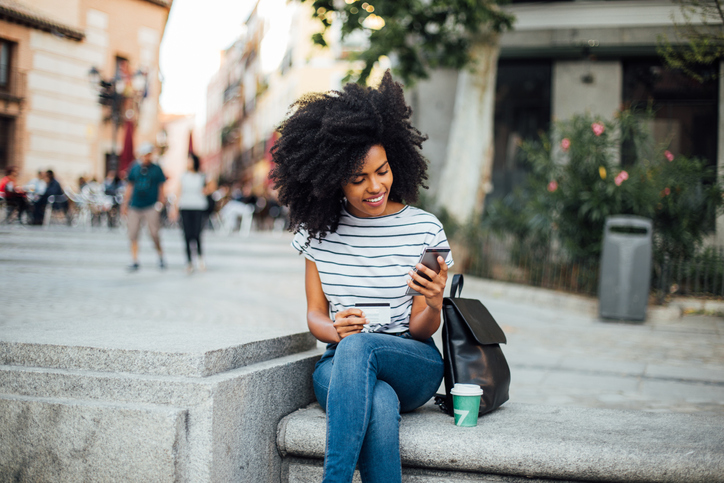 Beautiful dark haired young woman sitting on some steps with coffee and her cell phone. 