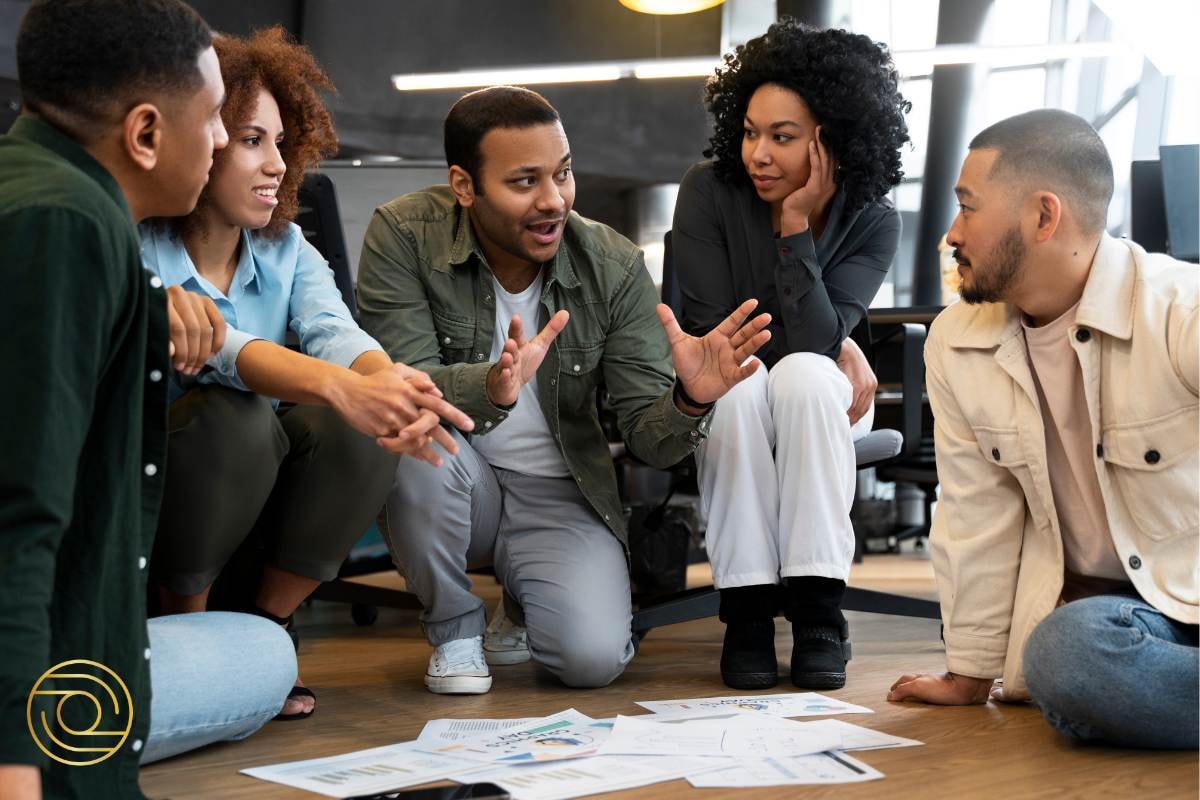 Team brainstorming session on the floor, with a leader explaining ideas while others listen attentively, papers spread around.