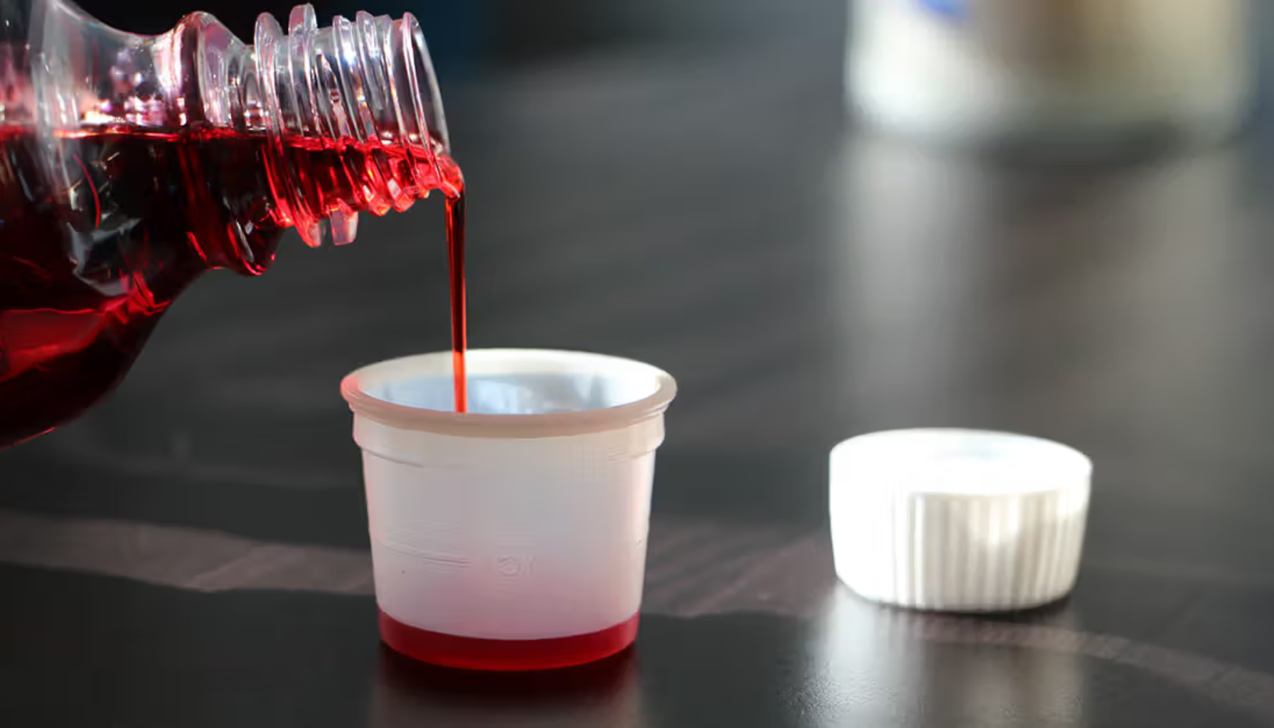 Rød flytende medisin helles fra flaske ned i et lite plastbeger for korrekt dosering.